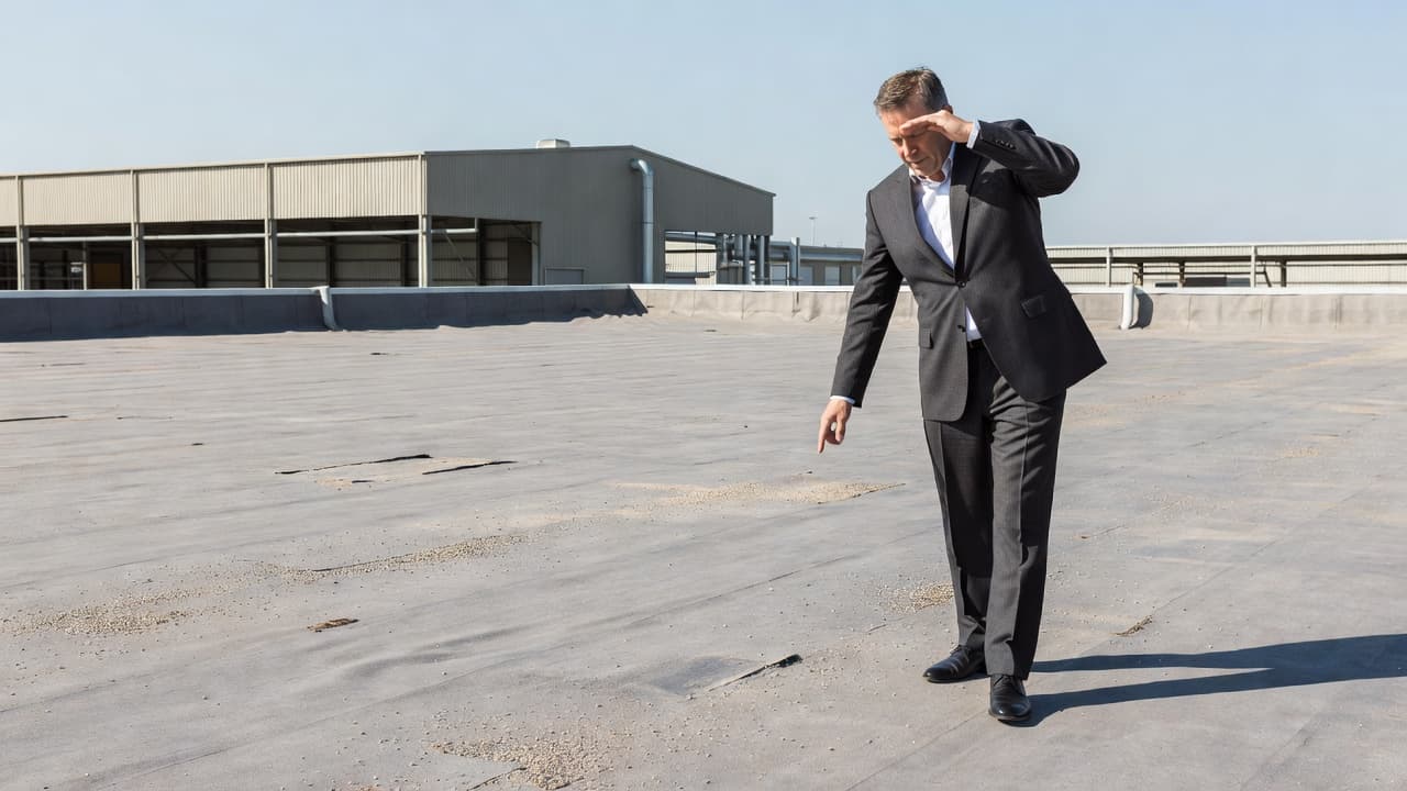 Can Exposed Nails Cause Roof Leaks After High Winds? A professional in business attire walks across a flat industrial rooftop while examining the roofing membrane under bright sunlight. The wide angle shot captures the vast, weathered surface of the facility.