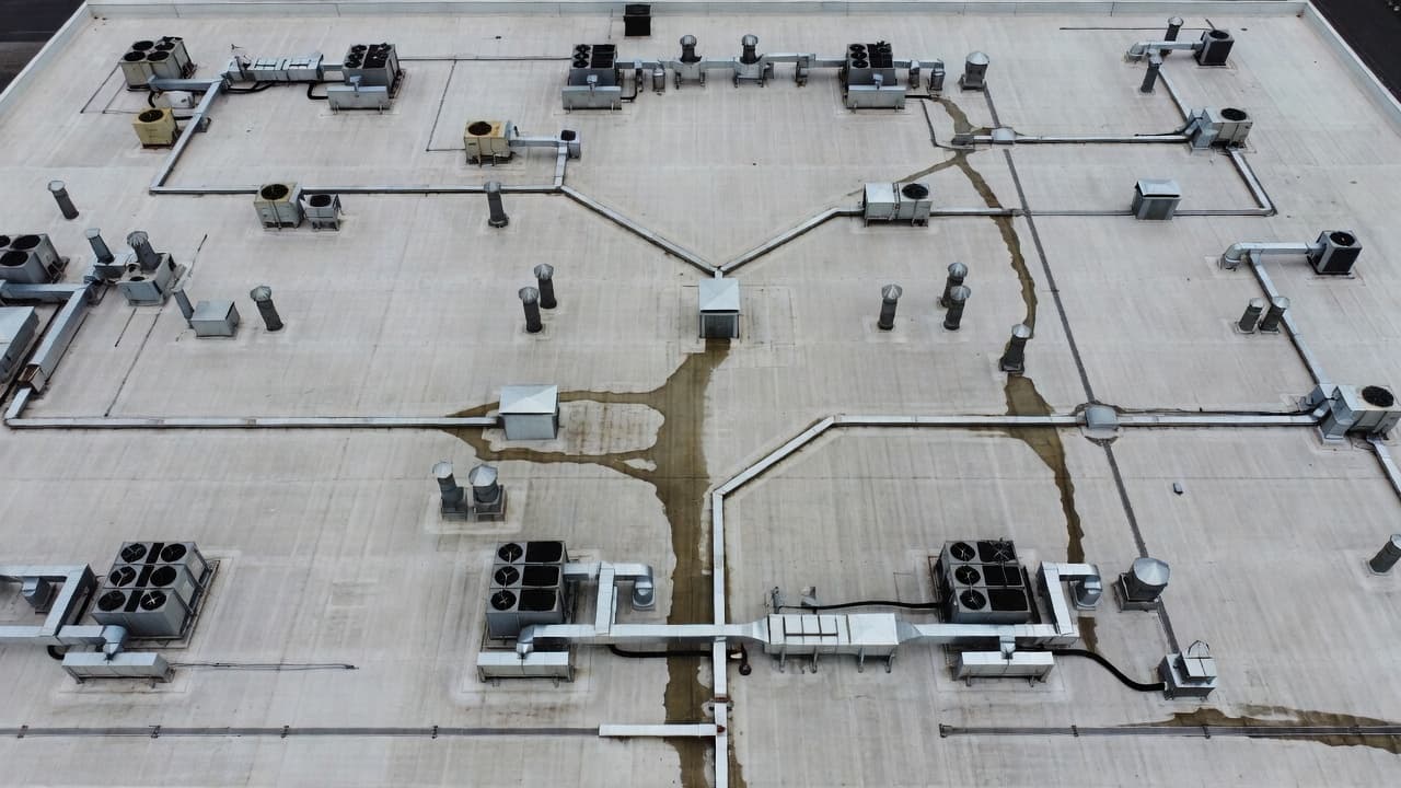 Can a Roof Leak Show Up Far From the Damage? A top-down aerial view displays a wide industrial flat roof filled with metal HVAC units, ventilation pipes, and complex drainage systems. The grey surface shows signs of wear on a cloudy day.