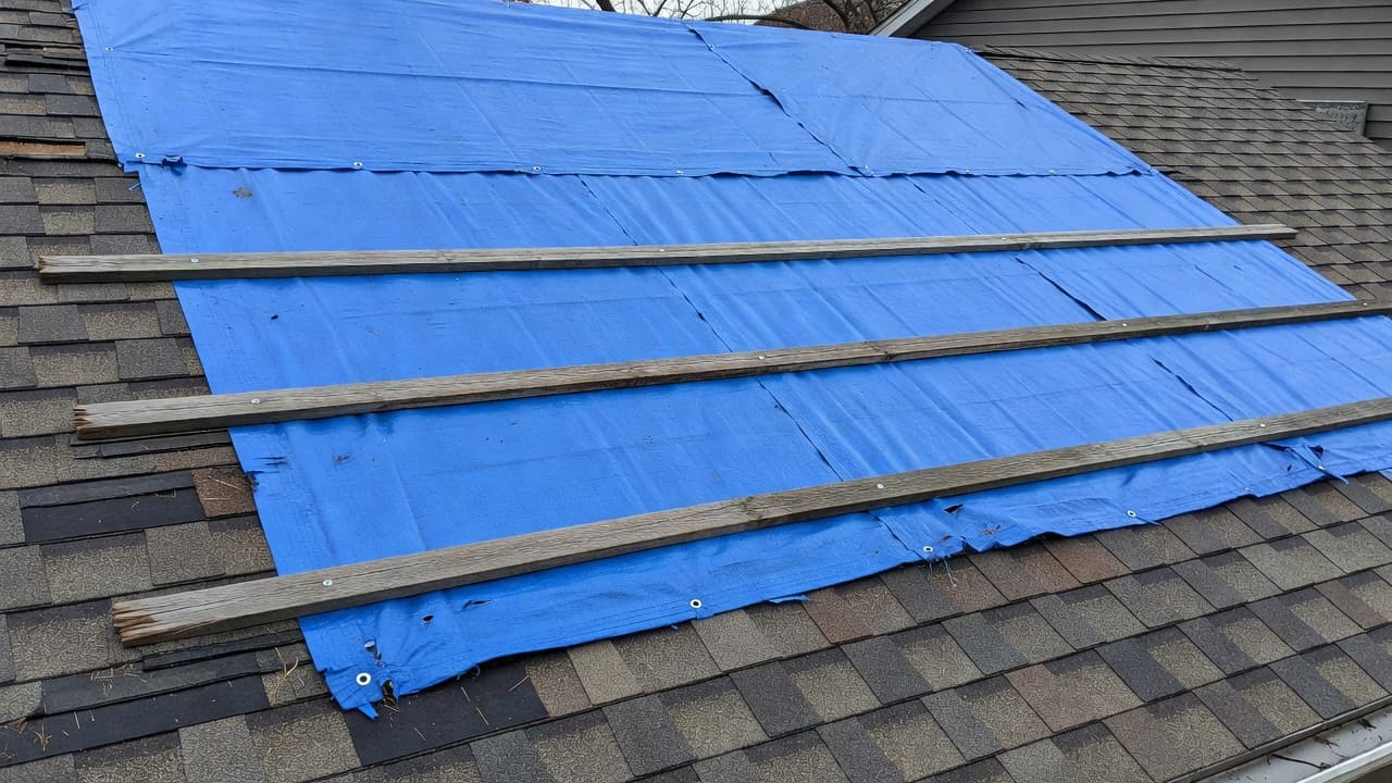 How Long Can a Roof Tarp Stay Up in Minnesota? A durable blue tarp is tightly secured across a damaged residential asphalt roof using wooden battens and metal screws. Visible wear indicates long-term exposure to harsh weather conditions under overcast skies.