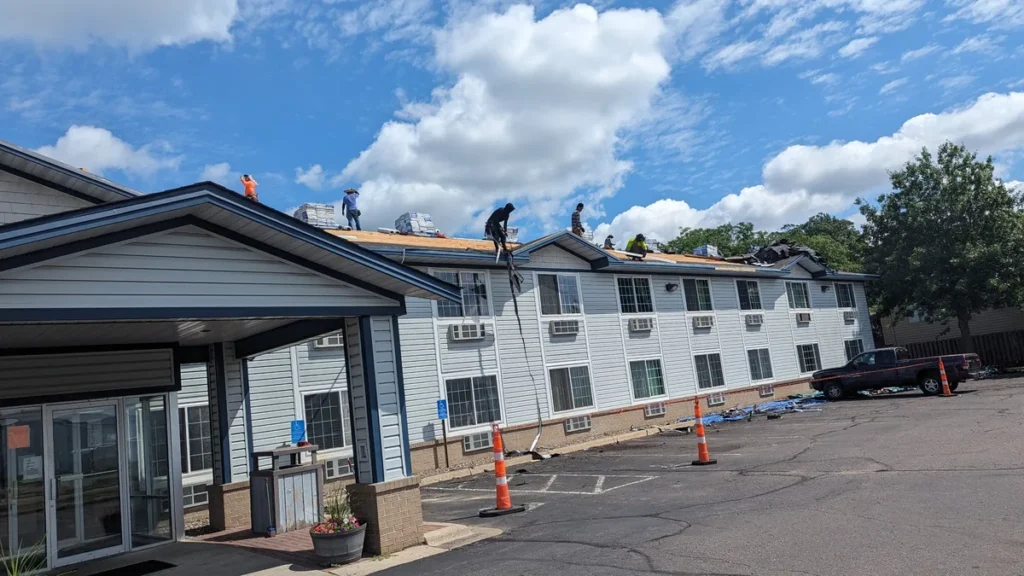 Roof replacement in progress Roofing crew replacing shingles on a two-story building, with materials on the roof and traffic cones below.