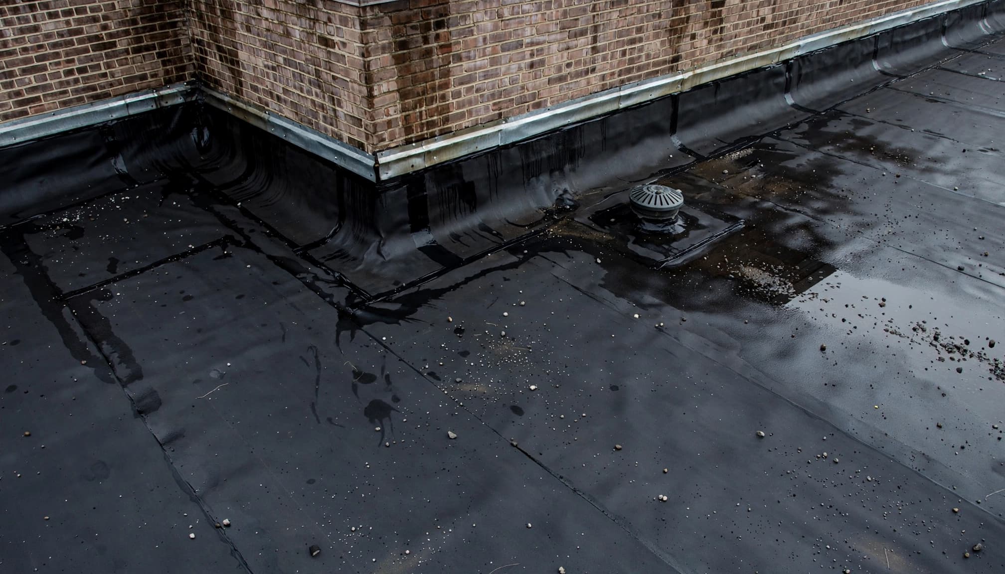 Can Wind-Driven Rain Enter Through Roof-to-Wall Flashing in St. Paul? A damp EPDM roof membrane turns up against a masonry wall beside a roof drain.